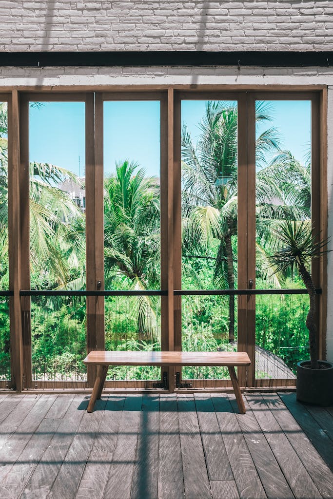 Wooden chair placed in spacious villa near panoramic windows overlooking lush green tropical garden against cloudless blue sky